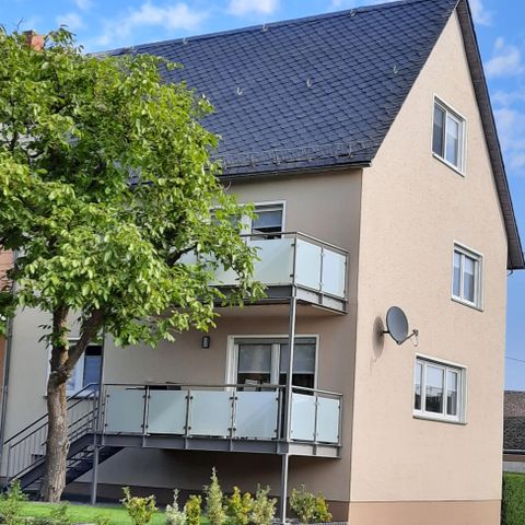 Ein modernes Wohnhaus mit einem Balkon und einem großen Baum im Vordergrund. Die Fassade ist hell und es gibt einen klaren blauen Himmel.