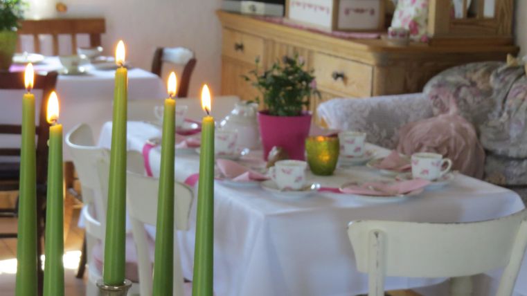 Ein gemütlicher Essbereich mit einem Tisch, der mit weißem Tischtuch, Geschirr und Pflanzen dekoriert ist. Grüne Kerzen brennen und verleihen der Szene eine einladende Atmosphäre.