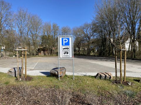 Ein Parkplatz mit einem Schild, das auf Parkmöglichkeiten hinweist. Rund um den Parkplatz stehen einige junge Bäume und Steine.