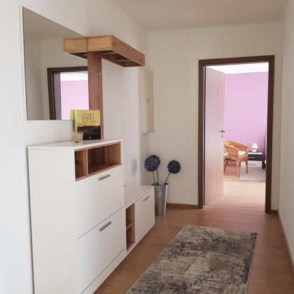 Ein heller Flur mit einem modernen Schuhschrank und einem Spiegel. Im Hintergrund ist ein Zugang zu einem Raum mit einem lila Wandfarbe und einem gemütlichen Sessel sichtbar.