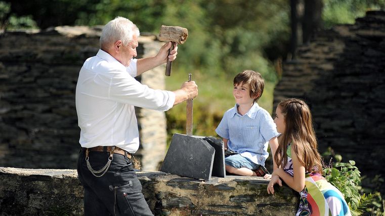 Ein älterer Mann zeigt zwei Kinder ein handwerkliches Werkzeug. Sie sitzen auf einer Mauer, umgeben von einer naturnahen Umgebung.