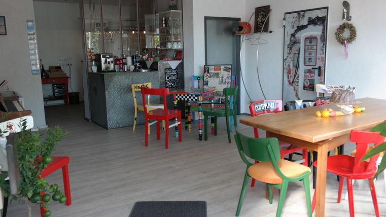 Ein helles Café mit bunten Stühlen und einem großen Holztisch. Der Raum ist modern und einladend gestaltet.