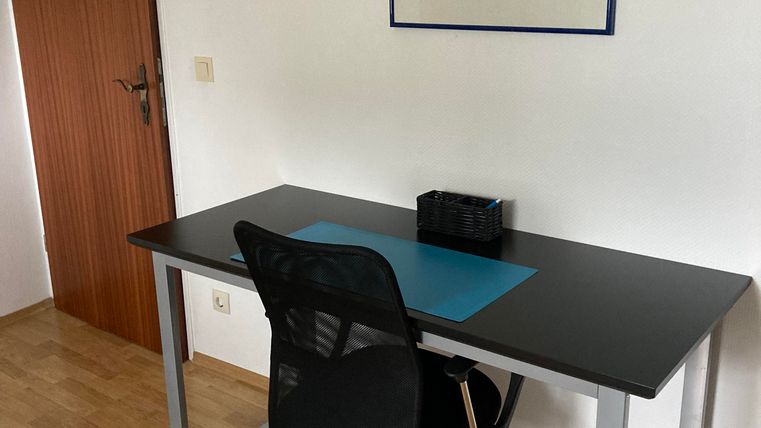 Ein einfacher Arbeitsplatz mit einem schwarzen Tisch und einem Bürostuhl. An der Wand hängt ein Bild von bunten Blumen.