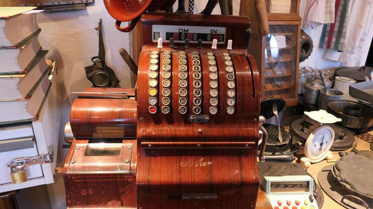 Eine alte Registerkasse aus Holz mit vielen Tasten. Sie steht auf einem Tisch umgeben von verschiedenen Gegenständen und Büchern.