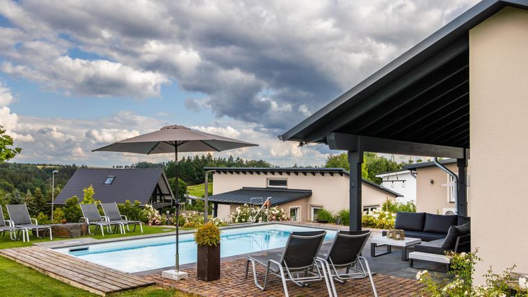 Ein modernes Haus mit einer Terrasse und einem Schwimmbad. Im Hintergrund sind grüne Hügel und Wolken am Himmel zu sehen.