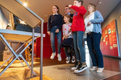 Eine Gruppe von fünf Personen springt gemeinsam in einem Ausstellungsraum. Im Hintergrund sind farbige Wände und ein Ausstellungsobjekt zu sehen.