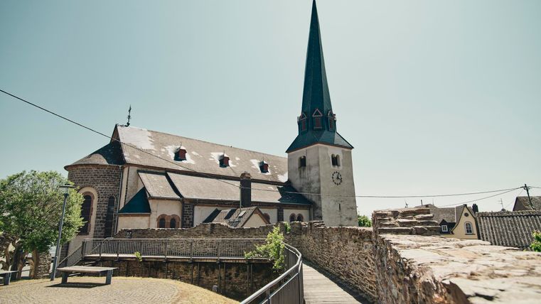 Eine alte Kirche mit einem hohen, spitzen Turm steht neben einer Mauer. Die Umgebung ist ruhig und sonnig.
