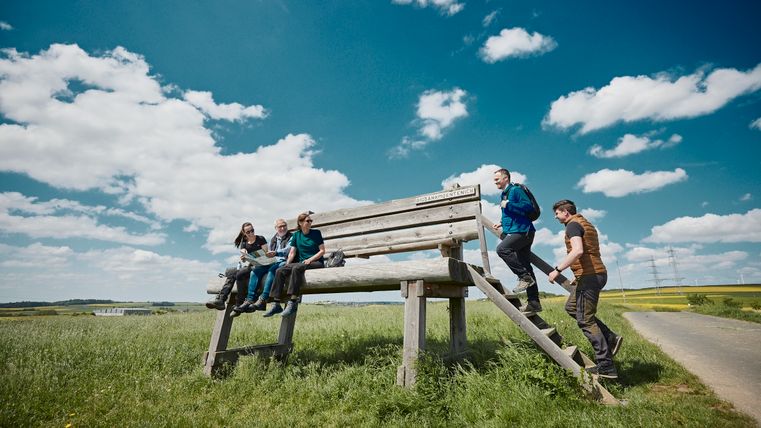 Die XXL-Bank auf einer grünen Wiese in Möntenich. Drei Menschen sitzen bereits auf der Bank und zwei weitere steigen über die Treppe auf die Bank.
