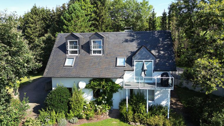 Ein schönes Haus mit einem grauen Dach und einem Balkon. Umgeben von gepflegten Gärten und Bäumen.