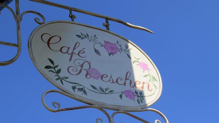 Ein Schild des Cafés „Roeschen“ hängt vor einem klaren blauen Himmel. Es ist floral gestaltet und zeigt den Namen in eleganten Buchstaben.