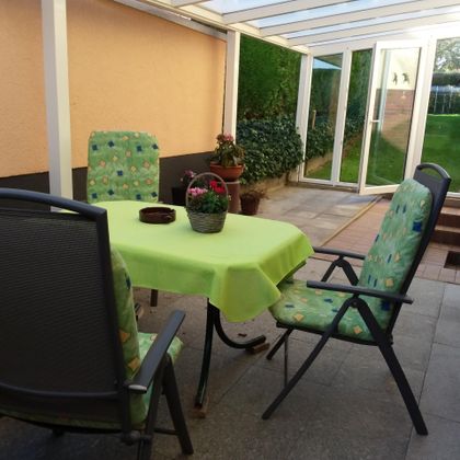 Ein gemütlicher Sitzbereich im Freien mit einem grünen Tisch und Stühlen. Vor dem Fenster steht ein Topf mit Blumen.
