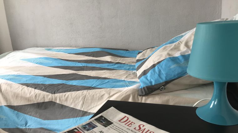 Ein gemütliches Schlafzimmer mit einem Bett und einer modernen Bettdecke in Blau- und Grautönen. Auf dem Nachttisch liegt eine Zeitung und eine stilvolle Tischlampe.