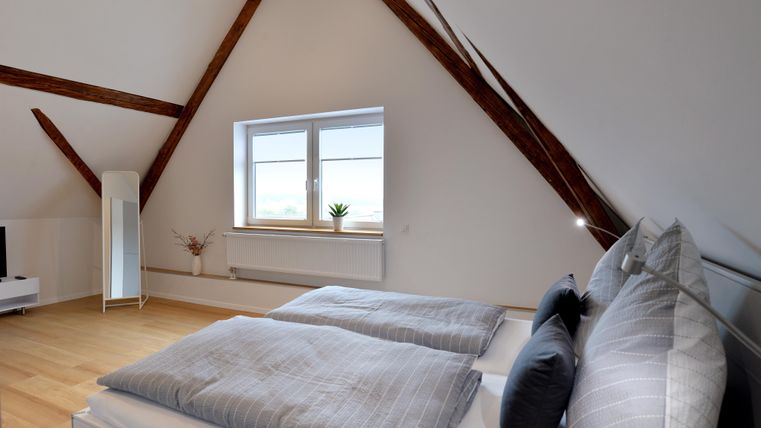 Ein modernes Schlafzimmer mit schrägen Deckenbalken und einem großen Fenster. Es ist hell und einladend, mit einem gemütlichen Doppelbett und minimalistischer Einrichtung.