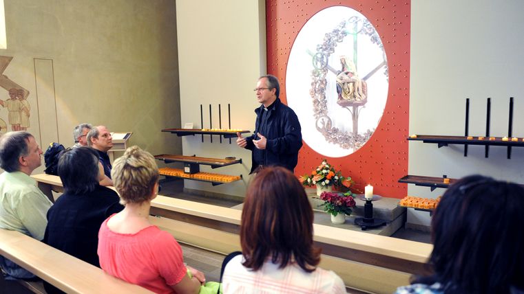 Heller Innenraum einer modernen Kirche. Personen sitzen auf einer der Holzbänke, im Vordergrund ist ein Pfarrer zu sehen.