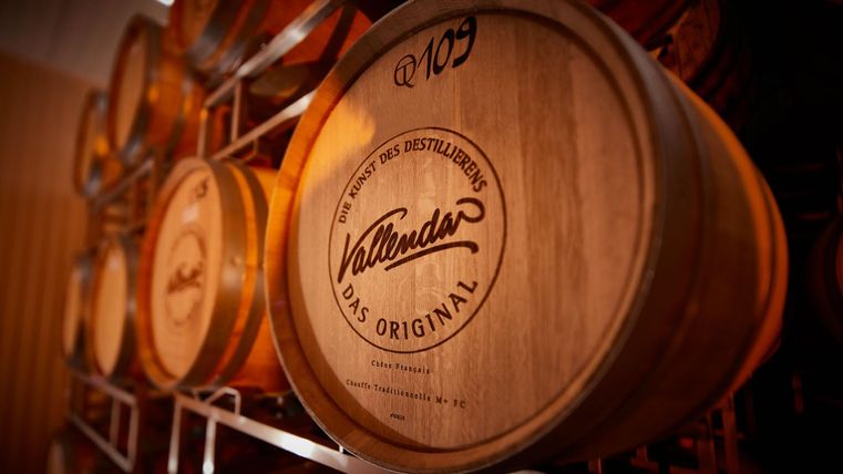 Een rek systeem met talloze houten vaten die worden gebruikt voor de opslag van sterke drank. Elk vat heeft een label met de tekst "Vallendar - Het Origineel".