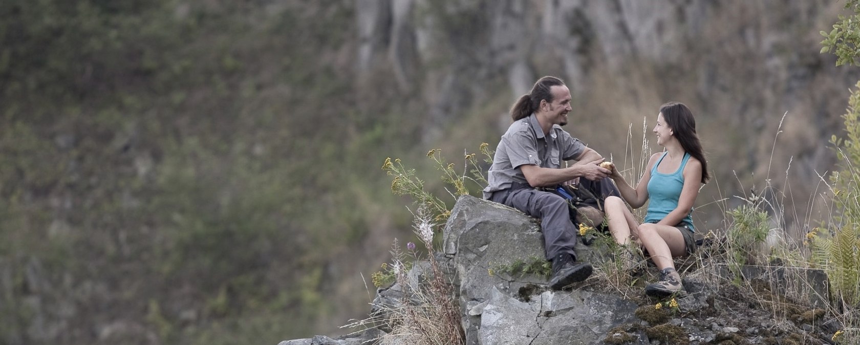 Zwei Personen sitzen auf einem Felsen im Steinbruch, umgeben von Pflanzen. Sie unterhalten sich und wirken entspannt., © Traumpfade/Kappest