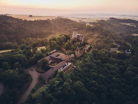 Eine beeindruckende Burg auf einem Hügel, umgeben von dichten Wäldern. Der Sonnenuntergang verleiht der Szene ein warmes Licht.