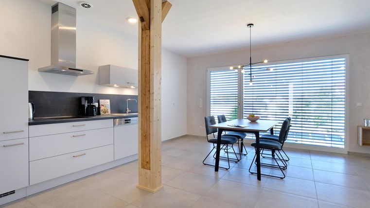 Eine moderne Küche mit einem Holzbalken und einem Esstisch für vier Personen. Große Fenster lassen viel Licht herein.