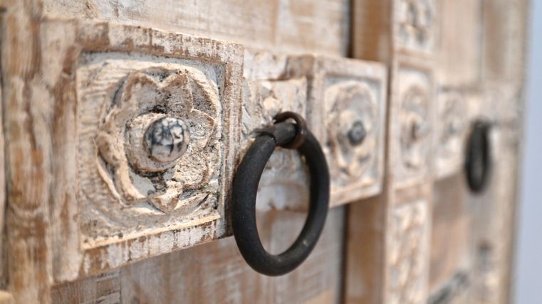 Ein dekoratives Holztürdetail mit einer Blumenmuster-Aussparung und einem runden Ringgriff. Die Oberfläche ist in einem rustikalen, weißen Farbton gehalten.