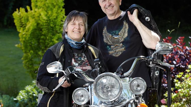 Ein Paar steht neben ihren Motorrädern in einem blühenden Garten. Beide tragen Motorradkleidung und lächeln glücklich in die Kamera.