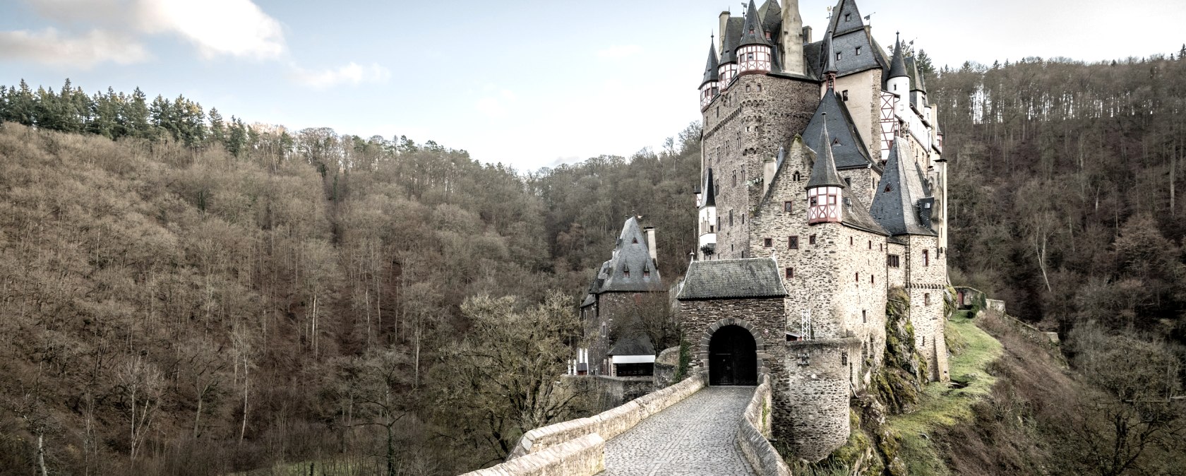 Burg Eltz thront majestätisch auf einem Hügel, umgeben von Wald. Ein gepflasterter Weg führt zur Burg, die von Türmen und Zinnen geprägt ist., © Eifel Tourismus GmbH, Dominik Ketz