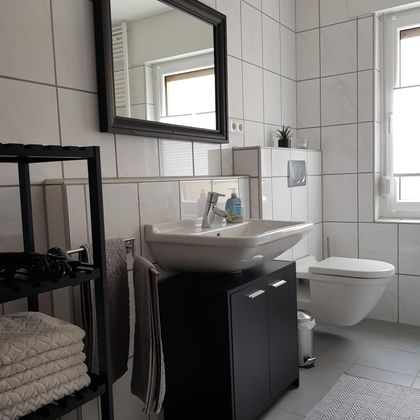 Ein modernes Badezimmer mit weißen Fliesen und einer großen Spiegelfläche. Es gibt ein Waschbecken, eine Toilette und einen offenen Regal mit Handtüchern.