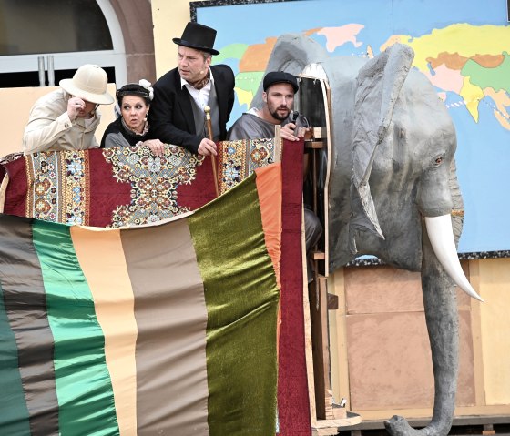 Darsteller des Theaterverein Kaisersesch spielen eine Szene aus &bdquo;In 80 Tagen um die Welt&ldquo;: Vier Figuren in historischen Kost&uuml;men blicken gespannt &uuml;ber einen bunt beh&auml;ngten Korb, der einen Hei&szlig;luftballon darstellt, w&auml;hrend neben ihnen ein gro&szlig;er Elefantenkopf als B&uuml;hnenrequisite und eine Weltkarte im Hintergrund zu sehen sind.