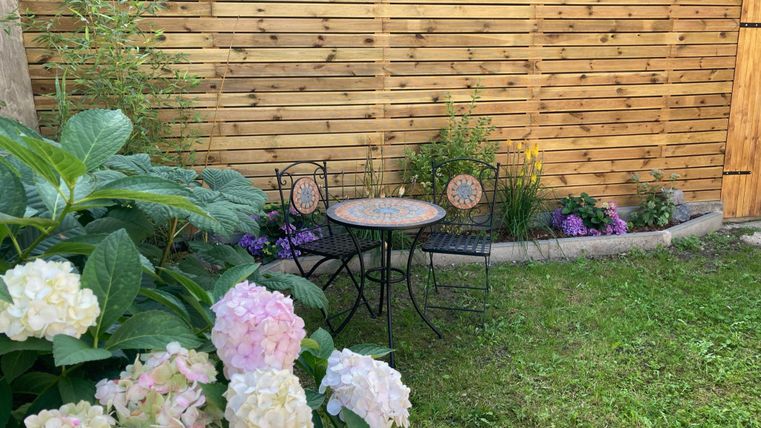 Ein schöner Garten mit einem kleinen Tisch und zwei Stühlen vor einer Holzwand. Bunte Blumen und grüne Pflanzen umgeben den Sitzbereich.