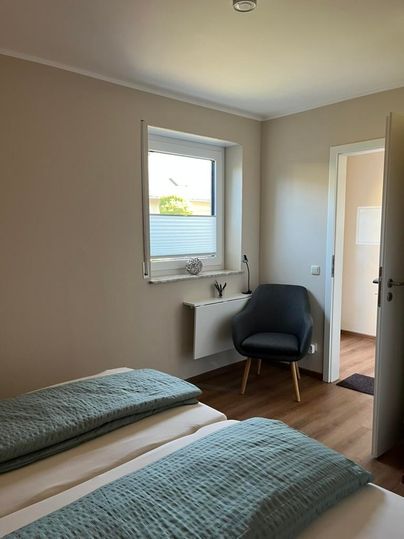 Ein helles Schlafzimmer mit zwei Betten und einer gemütlichen Ecke zum Sitzen. Der Raum hat eine große Fensterfläche und einen modernen Stil.