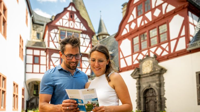 Ein Paar schaut sich eine Broschüre an, während sie in einer malerischen Altstadt stehen. Im Hintergrund sind schöne Fachwerkhäuser und eine klare Himmel zu sehen.