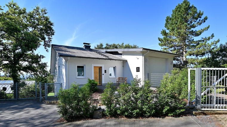 Ein modernes Einfamilienhaus mit einem weißen Außenanstrich und einem kleinen Vorgarten. Im Hintergrund stehen einige Bäume und der Himmel ist klar und blau.