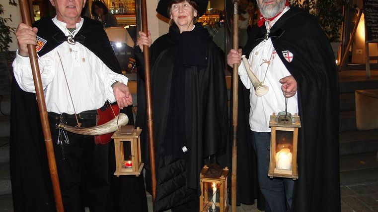 Drei Personen in historischen Kostümen halten Laternen und Stäbe. Sie stehen im Eingangsbereich eines Gebäudes und haben einen festlichen Ausdruck.