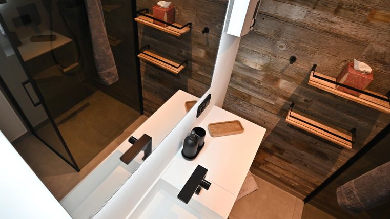 Ein modernes Badezimmer mit einem weißen Waschbecken und schwarzen Armaturen. Die Wände sind aus Holz, und es gibt offene Regale für Handtücher und Pflegeprodukte.