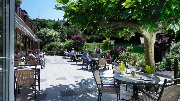 Eine einladende Terrasse mit Tischen und Stühlen im Freien. Im Hintergrund sind gepflegte Gärten und Gäste, die die Sonne genießen.
