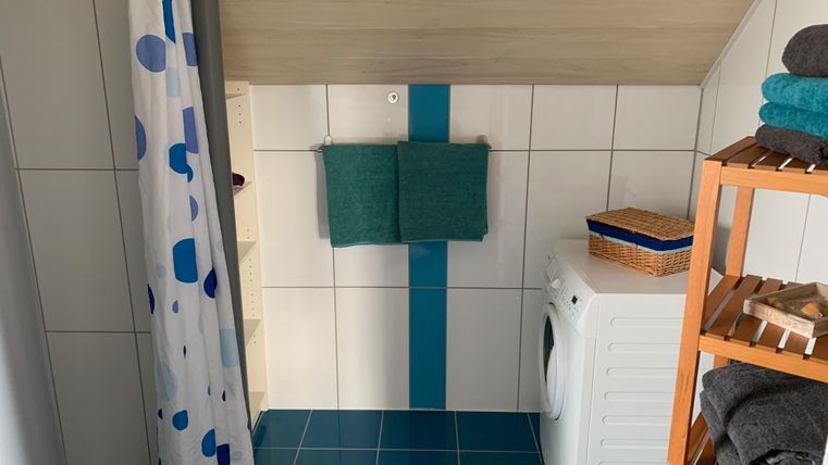 Een badkamer met blauwe tegels en een douche. Op een plank zijn handdoeken en een wasmand zichtbaar.