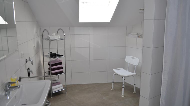 Ein modernes Badezimmer mit weißen Fliesen und einem Dachfenster. Es gibt eine Waschgelegenheit, einen Stuhl und einen Handtuchständer.