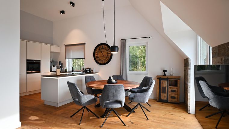 Ein modernes Esszimmer mit einem runden Tisch und bequemen Stühlen. Die Einrichtung ist hell und einladend, mit großen Fenstern, die Tageslicht hereinlassen.