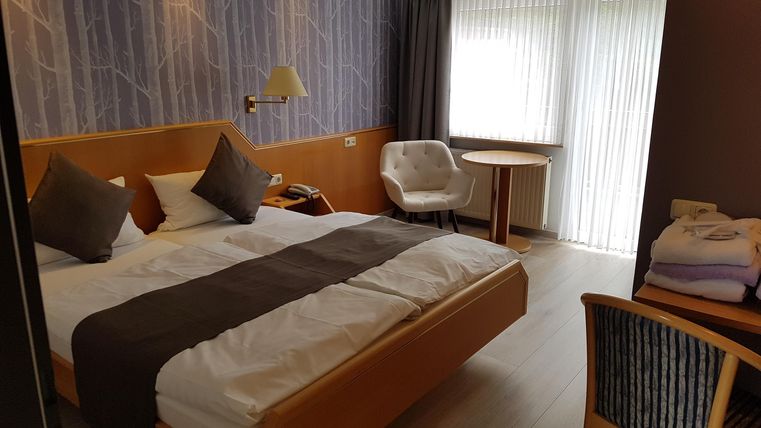 Ein modernes Hotelzimmer mit einem Doppelbett und zwei gemütlichen Stühlen. Das Zimmer hat große Fenster, die viel Tageslicht hereinlassen.