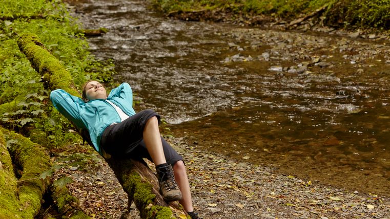 Wanderin lehnt entspannt an einem moosbedeckten Baumstamm am Bachufer.