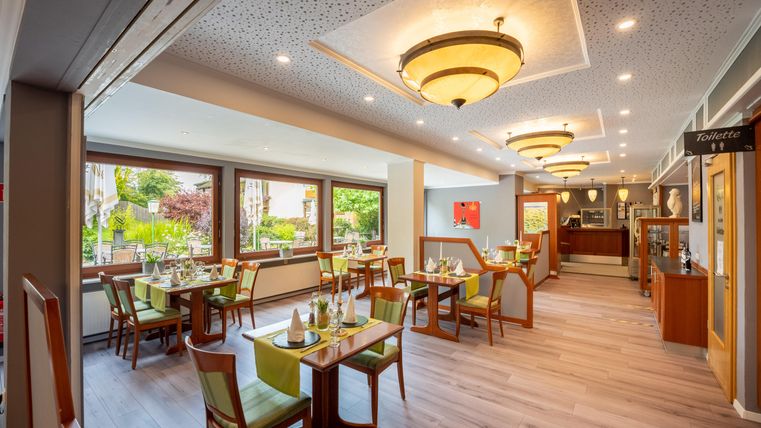 Ein elegantes Restaurant mit hellen Holzmöbeln und grünen Stühlen. Die großen Fenster bieten einen Blick auf einen gepflegten Garten.