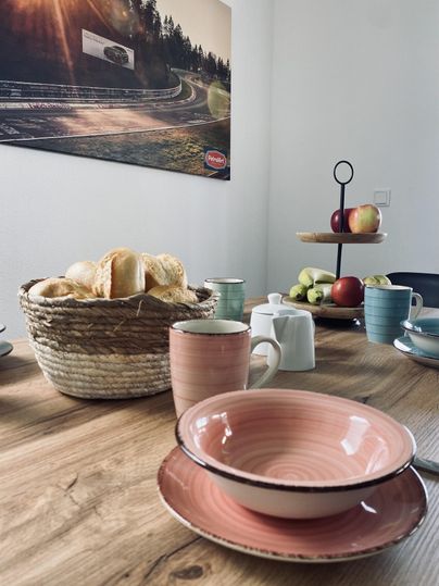 Ein schön gedeckter Tisch mit verschiedenen Tellern und Tassen. Im Hintergrund ist ein Korb mit Brötchen und eine Obstschale zu sehen.