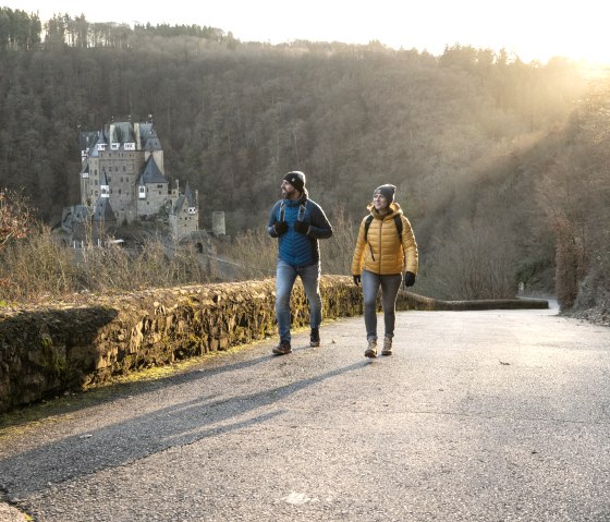 Zwei Wanderer auf einem Weg, die Burg Eltz im Hintergrund. Die Sonne scheint durch die Bäume, herbstliche Stimmung., © Eifel Tourismus GmbH, Dominik Ketz