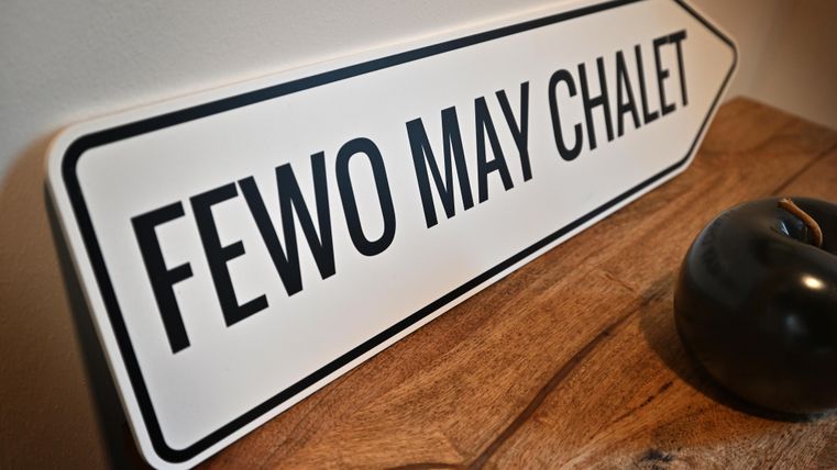 Ein Schild mit der Aufschrift "FEWO MAY CHALET" liegt auf einem Holztisch. Es hat ein schlichtes, modernes Design.