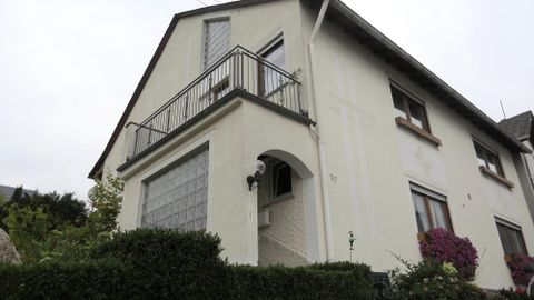 Ein weißes Haus mit einem Balkon und einem begrünten Vorgarten. Der Himmel ist bewölkt und die Atmosphäre wirkt ruhig.