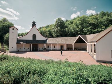 Zu sehen ist die Kloster und die Wallfahrtskirche Maria Martental. Die Helle Kapelle mit weißer Fassade und kleinem Zwiebelturm, umgeben von bewaldeten Hügeln. Vor dem Gebäude liegt ein großer, gepflasterter Innenhof mit überdachtem Kreuzgang und Sitzgelegenheiten unter freiem Himmel.