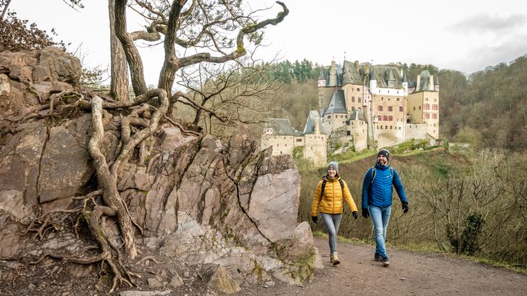 Zwei Wanderer auf einem Pfad mit Burg Eltz im Hintergrund.