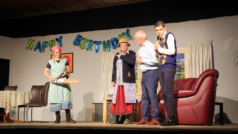 Mehrere Darstellerinnen und Darsteller auf einer Theaterbühne während einer humorvollen Geburtstagsszene mit dekorierter Wohnkulisse.