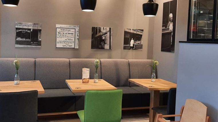 Ein modernes Café mit gemütlichen Tischen und bunten Stühlen. An der Wand hängen historische Fotografien, die eine einladende Atmosphäre schaffen.