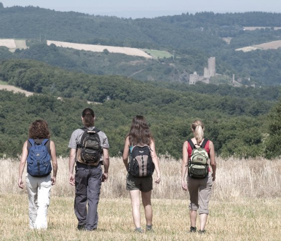 Vier Wanderer mit Rucksäcken gehen über ein Feld. Im Hintergrund ist die Burgruine Monreal zu sehen, umgeben von bewaldeten Hügeln., © Traumpfade/Kappest
