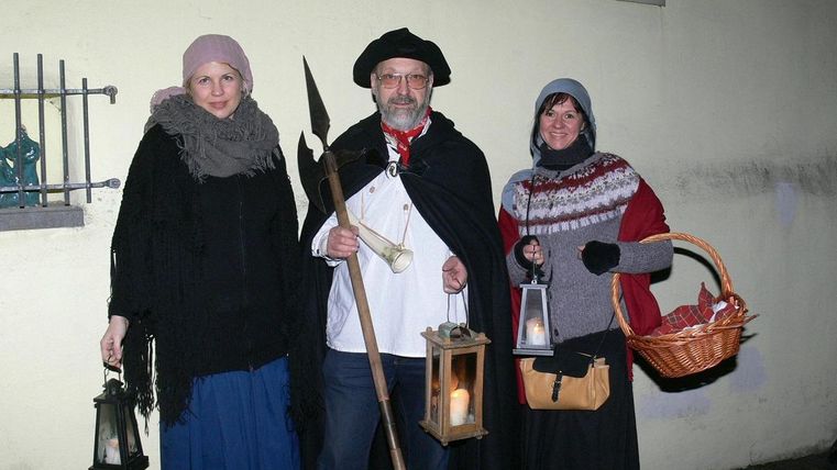 Drei Personen in historischen Kostümen stehen in der Nacht. Sie halten Laternen und andere Requisiten und posieren vor einer Wand.